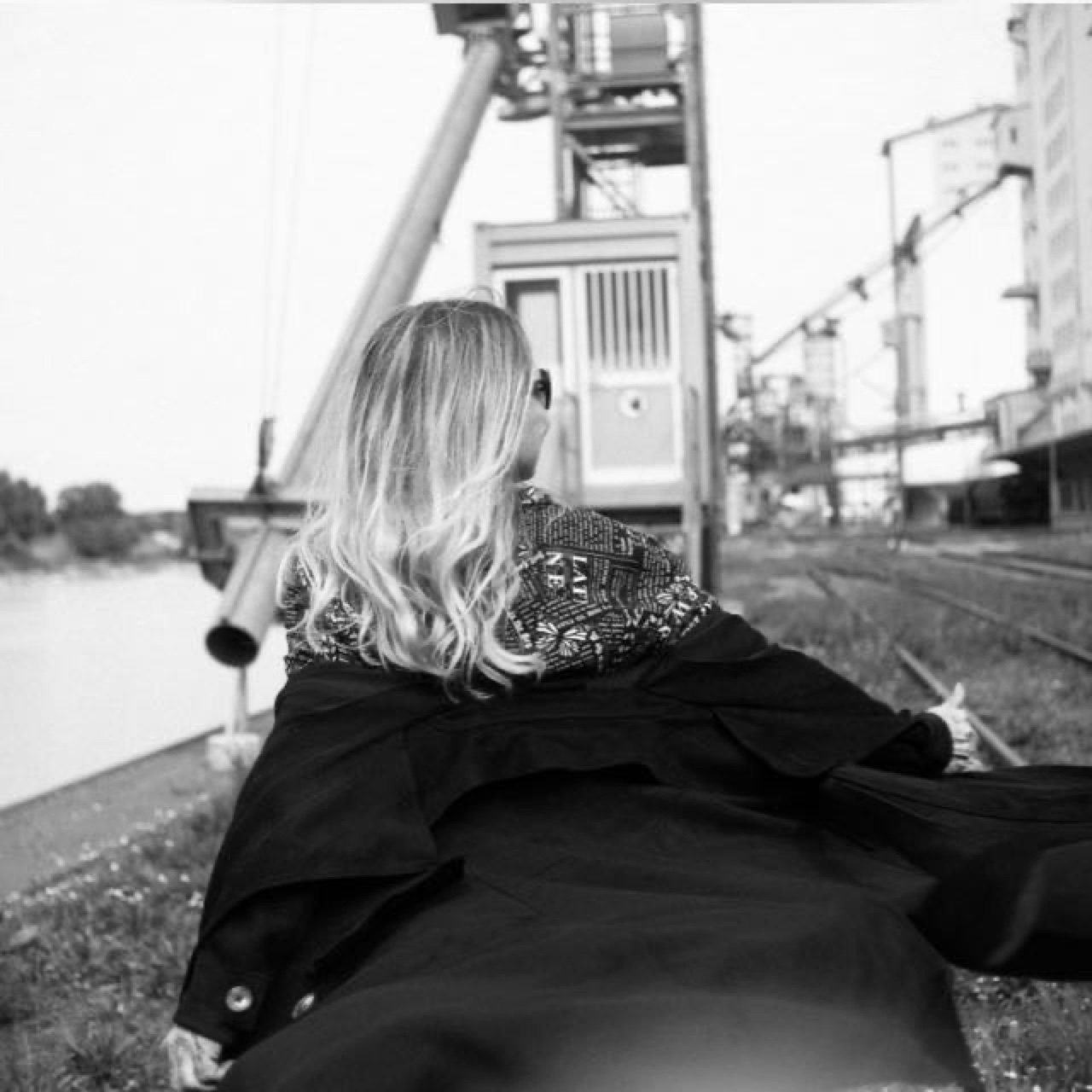 AS-T, the artist, seated by a river before an industrial harbour at dusk.